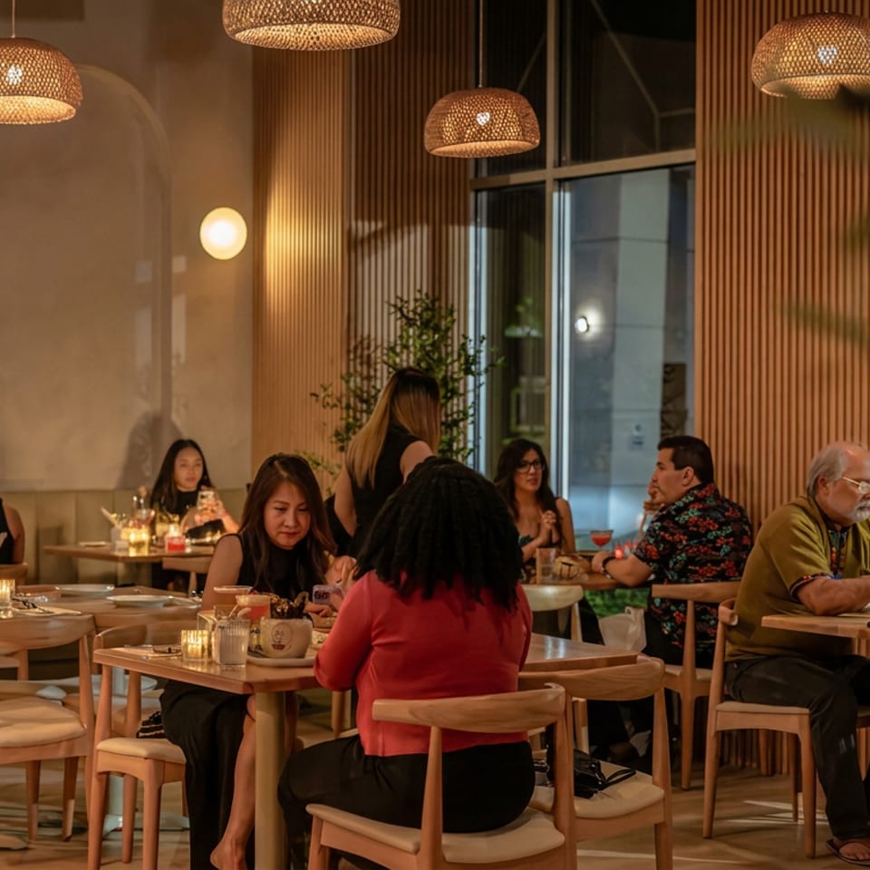 Warmly lit restaurant dining room with pendant lights, wooden furnishings, plants, and several groups of diners.