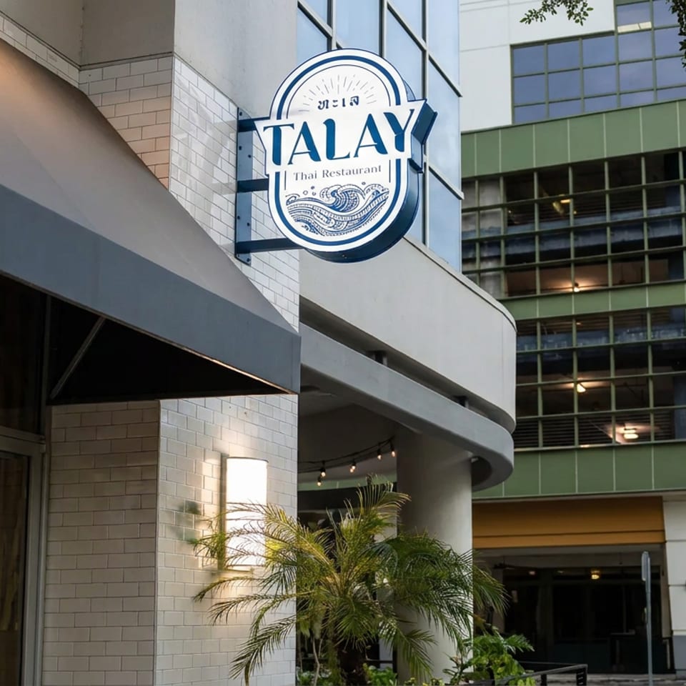 Exterior of Talay Thai Restaurant in a modern building, featuring prominent signage, palm plants, and a covered entrance.