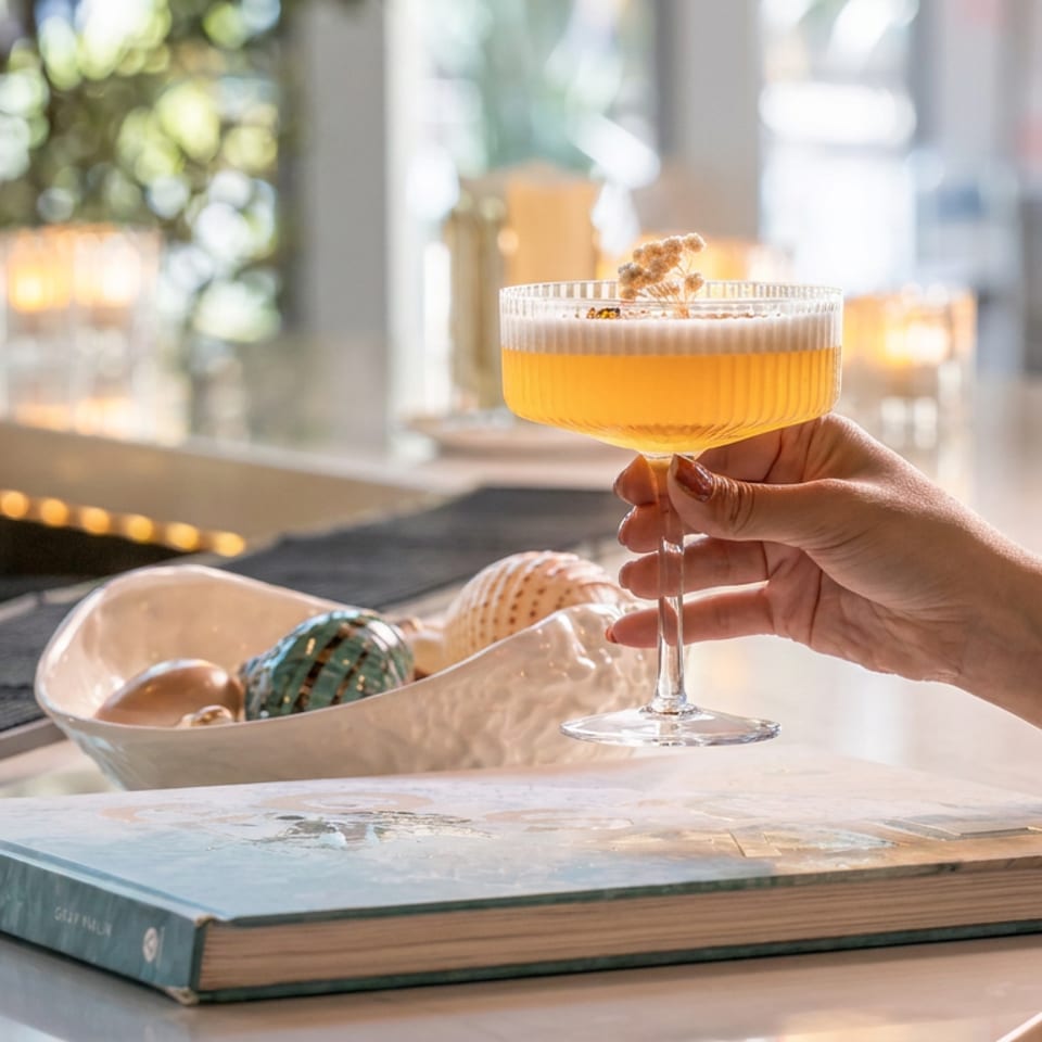 Hand holding a golden cocktail in a coupe glass at a sunlit bar, with a coastal book and decorative bowl nearby.