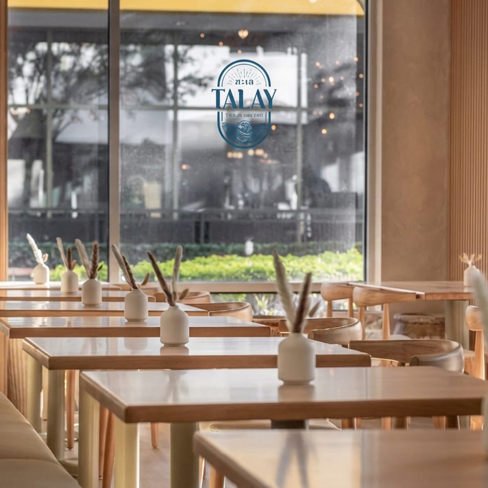 Warm, modern restaurant dining room with wooden tables, cream seating, decorative vases, and Talay logo on the window.