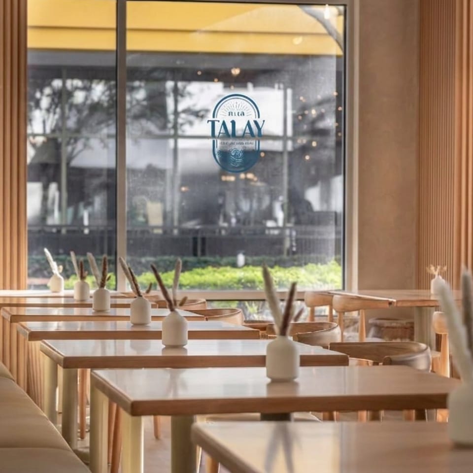 Sunlit Talay restaurant interior with wooden tables, white vase centerpieces, slatted walls, and booth seating.